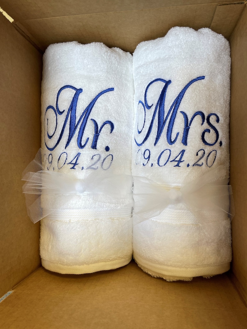 May include: Two white rolled towels featuring blue embroidered text. One towel reads Mr. 09.04.20 and the other reads Mrs. 09.04.20. Each towel is tied with a white tulle ribbon bow. The towels are positioned side by side inside a cardboard box.