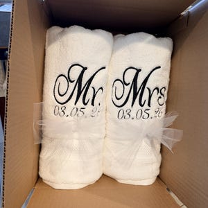 May include: Two white rolled towels featuring black embroidered text that reads Mr. and Mrs. above the date 03.05.26. Each towel is tied with a sheer white tulle ribbon and they are positioned side by side inside a brown cardboard shipping box.