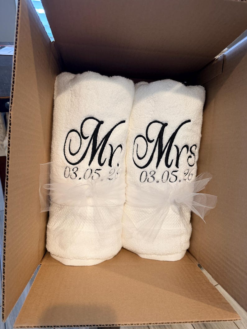 May include: Two white rolled towels featuring black embroidered text that reads Mr. and Mrs. above the date 03.05.26. Each towel is tied with a sheer white tulle ribbon and they are positioned side by side inside a brown cardboard shipping box.