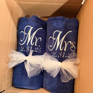 May include: Two navy blue towels rolled and tied with white tulle ribbons. The left towel is embroidered with white script text reading Mr. and the date 12.31.23. The right towel is embroidered with white script text reading Mrs. and the date 12.31.23.