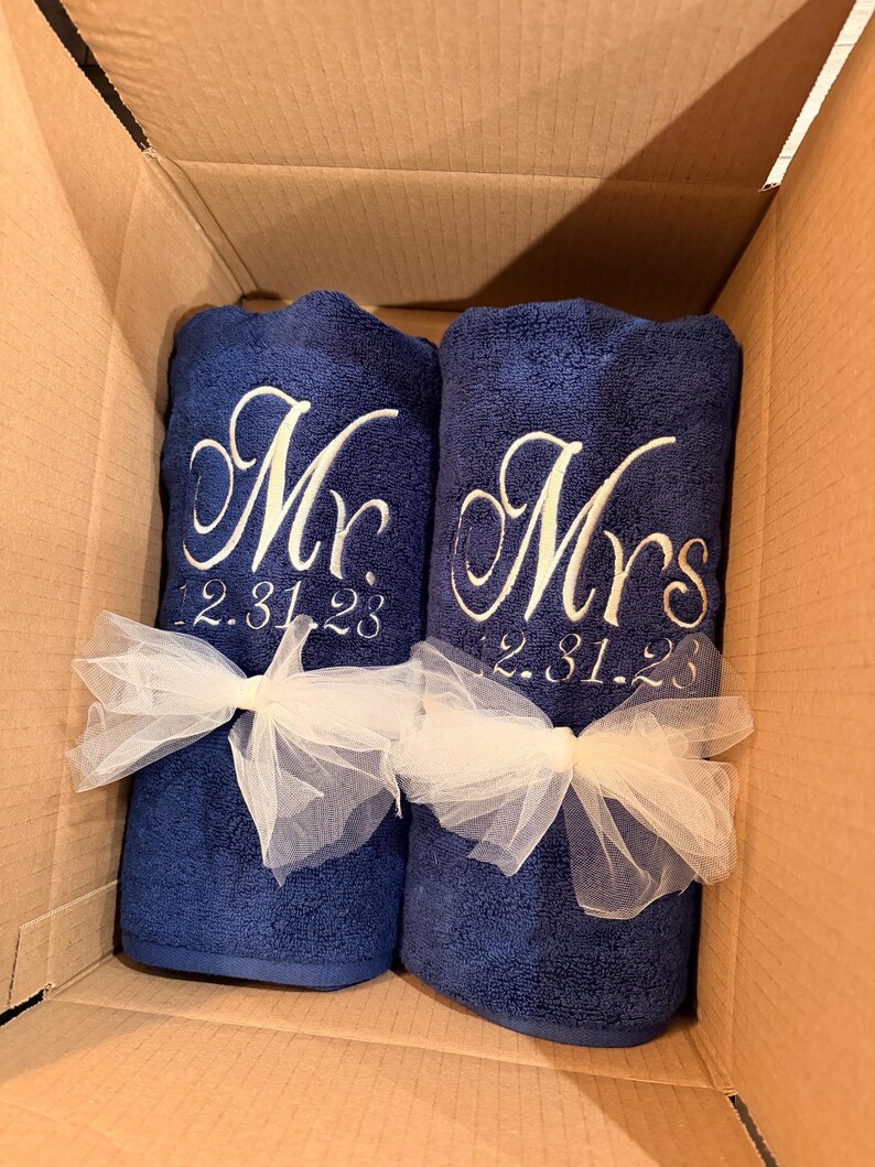 May include: Two navy blue towels rolled and tied with white tulle ribbons. The left towel is embroidered with white script text reading Mr. and the date 12.31.23. The right towel is embroidered with white script text reading Mrs. and the date 12.31.23.