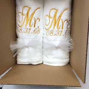May include: Two white towels rolled and placed inside a cardboard box. Each towel features gold embroidered text reading Mr. and Mrs. respectively, followed by the date 08.31.68. A white tulle ribbon is tied around the center of each towel.