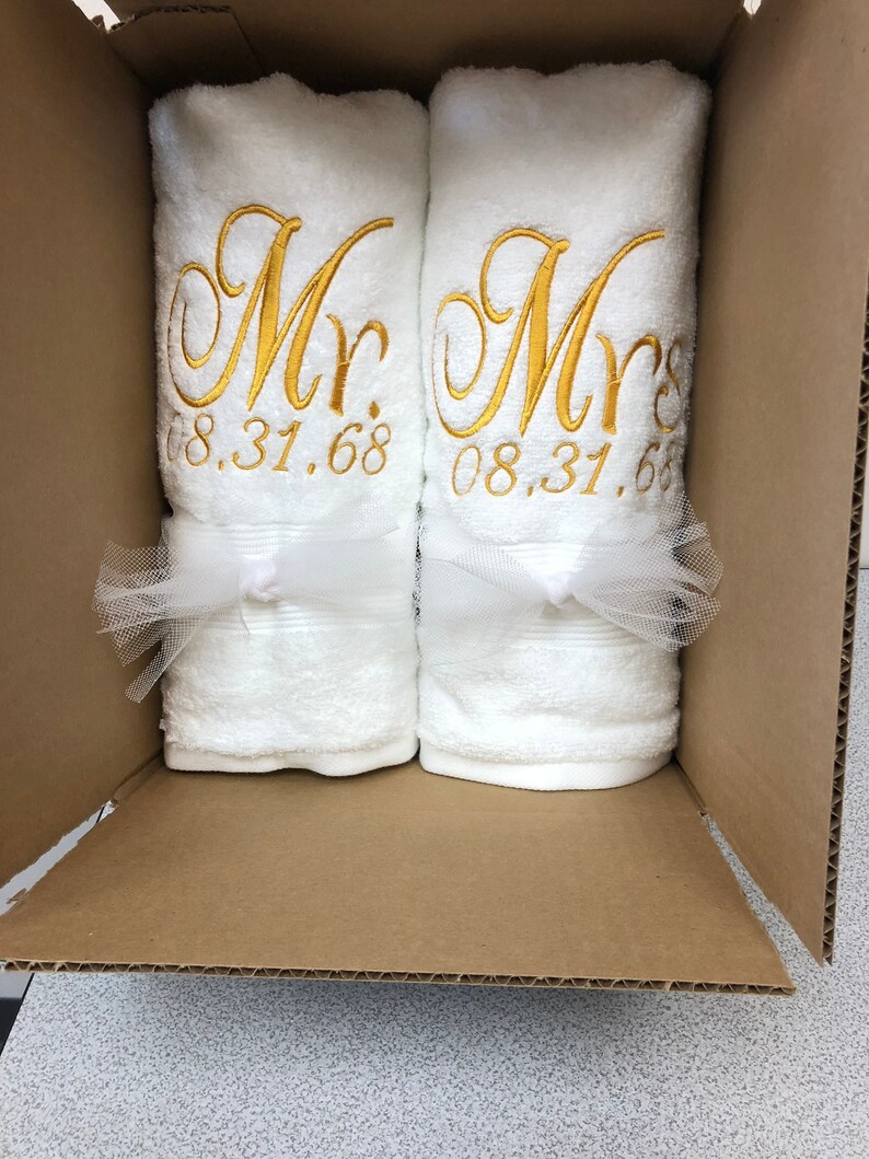 May include: Two white towels rolled and placed inside a cardboard box. Each towel features gold embroidered text reading Mr. and Mrs. respectively, followed by the date 08.31.68. A white tulle ribbon is tied around the center of each towel.