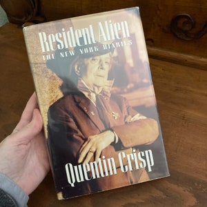 May include: A hardcover book titled "Resident Alien: The New York Diaries" by Quentin Crisp. The cover features a sepia-toned portrait of a man in a suit and hat. The book is held in a person's hand.