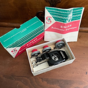 May include: A Singer Automatic Zigzagger sewing machine attachment set in a white box with a green and pink cardboard lid and instruction manual. The set includes a black metal attachment and three grey and red circular cams for stitching.