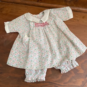 May include: A vintage baby outfit featuring a floral print in pink, green, and blue on a white background. The set includes a short-sleeved top with a white collar and a small pocket, paired with matching bloomers. The top has a decorative embroidered detail.