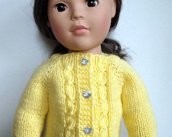 May include: A doll wearing a yellow knitted cardigan with cable knit detailing and three sparkly buttons. The doll has brown hair and is wearing black pants. The cardigan is a bright, cheerful yellow.