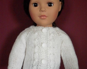 May include: A doll wearing a white, cable-knit sweater with three large, round white buttons. The sweater has a textured pattern and long sleeves. The doll has dark hair and is wearing black pants.