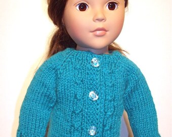 May include: A doll wearing a teal blue knitted cardigan with three clear, faceted buttons. The cardigan has a cable knit design and long sleeves. The doll has brown hair and brown eyes.