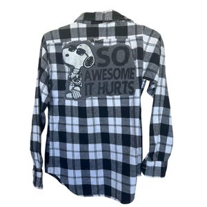 May include: Black and white plaid flannel shirt with a Snoopy graphic wearing sunglasses and the text "SO AWESOME IT HURTS". The shirt has long sleeves and a button-down front.