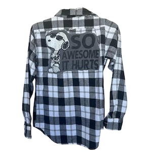 May include: Black and white plaid flannel shirt with long sleeves. The back of the shirt features a patch with Snoopy wearing sunglasses and the text "SO AWESOME IT HURTS."
