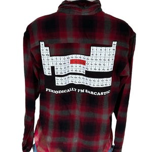 May include: Red and black plaid flannel shirt with a periodic table design on the back. The shirt features the text "PERIODICALLY I'M SARCASTIC" below the periodic table. The shirt has long sleeves and a button-down collar.