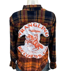 May include: A plaid flannel shirt in shades of orange, brown, and navy blue. The back features a white patch with the words "WRANGLING RECORDS" and an orange illustration of a cowboy on a horse.