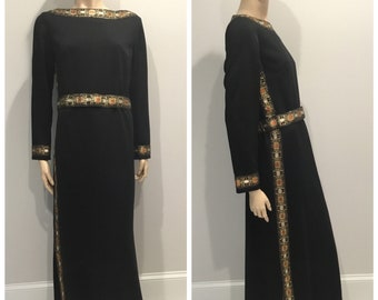 May include: A long black maxi dress with gold and orange geometric embroidery. The dress has a boat neck, long sleeves, and a side slit.
