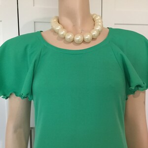 May include: A green short-sleeved dress with a round neckline and ruffled sleeves. The dress is worn by a mannequin with a large pearl necklace.