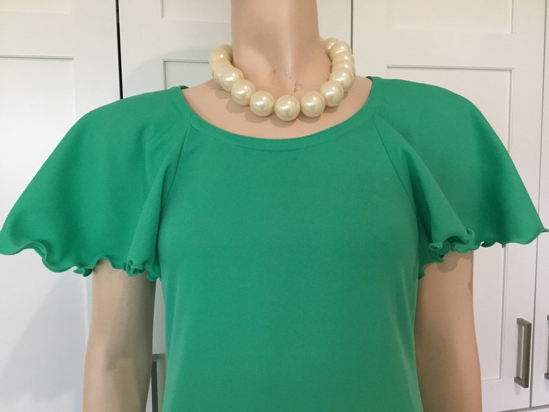May include: A green short-sleeved dress with a round neckline and ruffled sleeves. The dress is worn by a mannequin with a large pearl necklace.