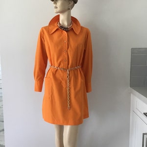 Op de afbeelding: Een feloranje getailleerde jurk met lange mouwen en &eacute;&eacute;n zak aan de linkerkant. De jurk heeft een gouden kettingriem en wordt gedragen door een mannequin met een bruin-oranje gestreepte hoed.