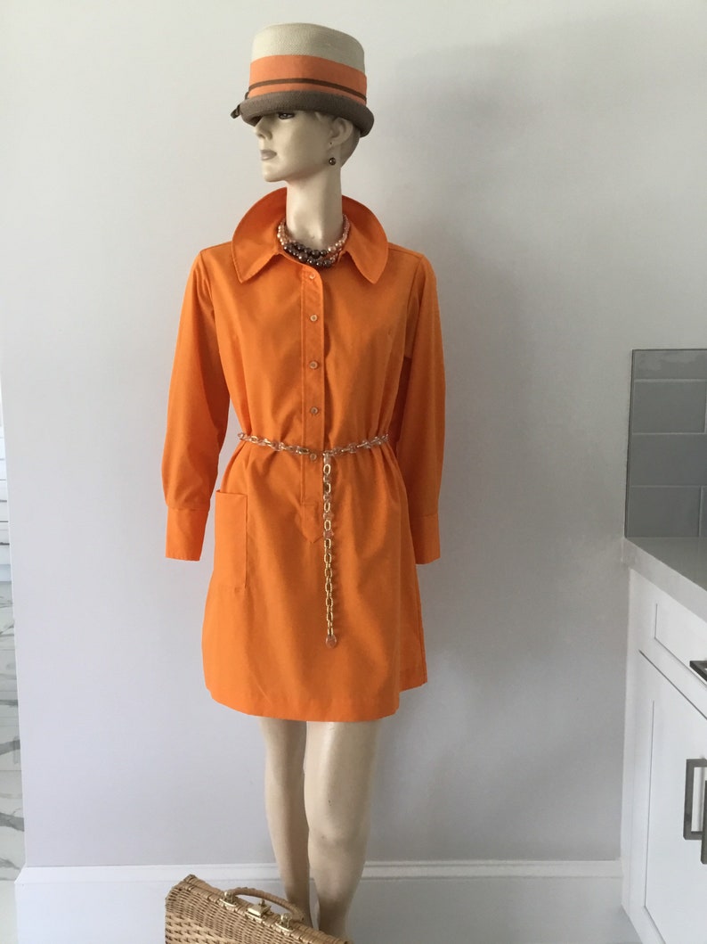 Op de afbeelding: Een feloranje getailleerde jurk met lange mouwen en &eacute;&eacute;n zak aan de linkerkant. De jurk heeft een gouden kettingriem en wordt gedragen door een mannequin met een bruin-oranje gestreepte hoed.