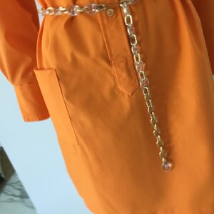 Op de afbeelding: Een feloranje shirtjurk met lange mouwen en een enkele zak aan de linkerkant. De jurk is in de taille getailleerd met een transparante plastic kettingriem.