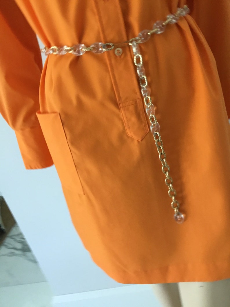 Op de afbeelding: Een feloranje shirtjurk met lange mouwen en een enkele zak aan de linkerkant. De jurk is in de taille getailleerd met een transparante plastic kettingriem.
