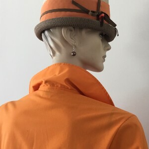 Op de afbeelding: Een bruine en beige strohoed met een bruin lint en een klein oranje accent. De hoed wordt gedragen door een mannequin met een oranje shirt.