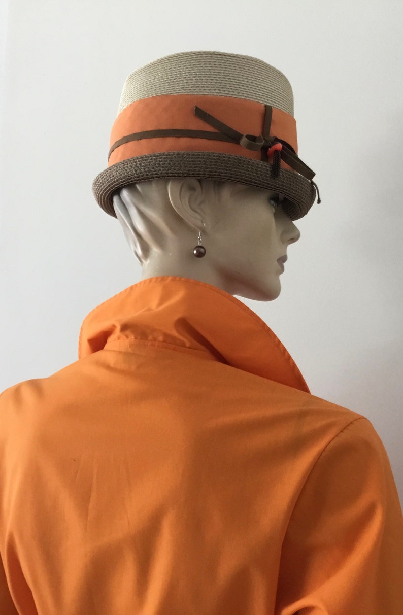 Op de afbeelding: Een bruine en beige strohoed met een bruin lint en een klein oranje accent. De hoed wordt gedragen door een mannequin met een oranje shirt.