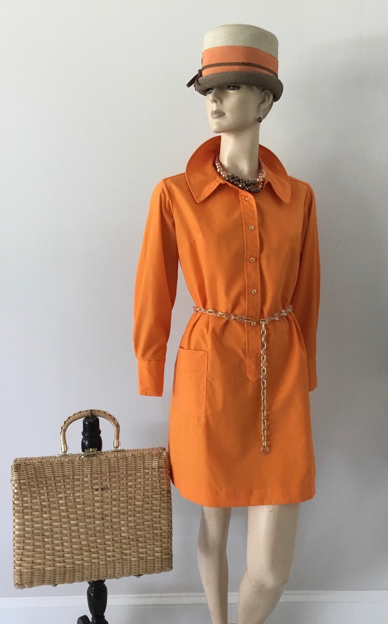 Op de afbeelding: Een feloranje mini-jurk met een kraag, lange mouwen en een knoopsluiting aan de voorkant. De jurk heeft een gouden kettingriem en een geweven strooien handtas.