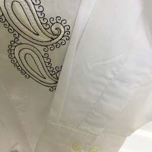 K&ouml;nnte beinhalten: Wei&szlig;es Langarmhemd mit schwarzer Paisley-Stickerei auf der linken Brustseite. Das Hemd hat einen Knopfkragenshirt und zwei Kn&ouml;pfe an der Manschette.