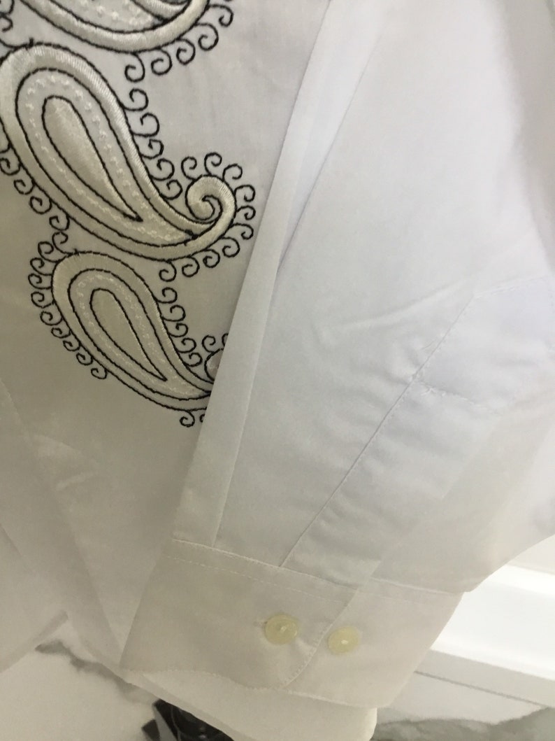 K&ouml;nnte beinhalten: Wei&szlig;es Langarmhemd mit schwarzer Paisley-Stickerei auf der linken Brustseite. Das Hemd hat einen Knopfkragenshirt und zwei Kn&ouml;pfe an der Manschette.