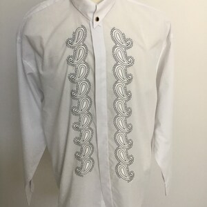 K&ouml;nnte beinhalten: Wei&szlig;es, lang&auml;rmliges Hemd mit Kn&ouml;pfen und einem Paisley-Muster auf der Vorderseite. Das Hemd hat einen Stehkragen und einen einzigen Knopfverschluss.