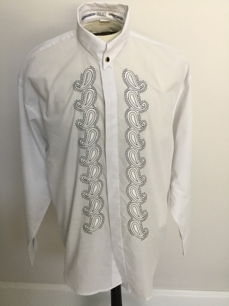 K&ouml;nnte beinhalten: Wei&szlig;es, lang&auml;rmliges Hemd mit Kn&ouml;pfen und einem Paisley-Muster auf der Vorderseite. Das Hemd hat einen Stehkragen und einen einzigen Knopfverschluss.