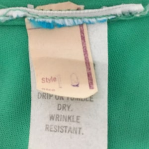 May include: A white tag with black text that reads "Style", "Drip or Tumble Dry", and "Wrinkle Resistant". The tag is attached to a green fabric.