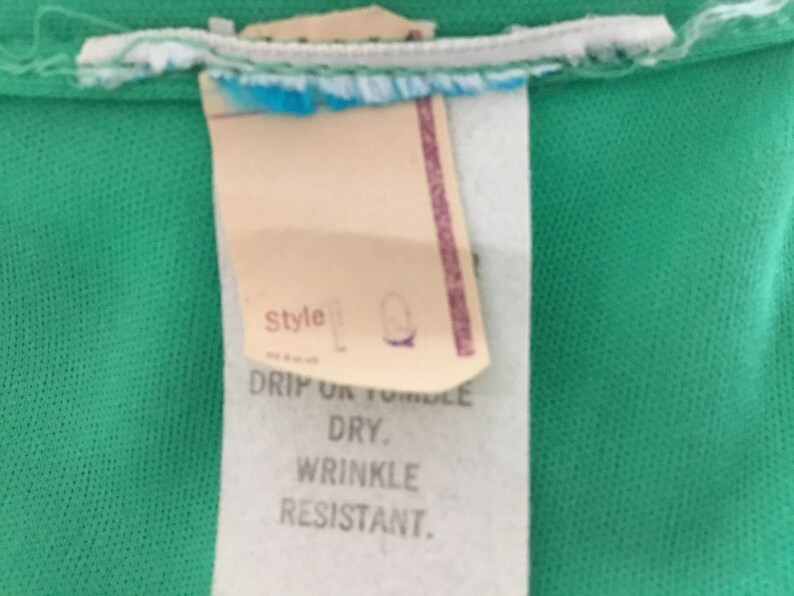 May include: A white tag with black text that reads "Style", "Drip or Tumble Dry", and "Wrinkle Resistant". The tag is attached to a green fabric.
