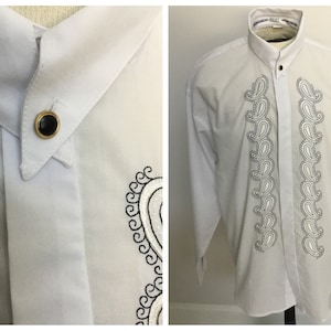 K&ouml;nnte beinhalten: Wei&szlig;es, lang&auml;rmliges Hemd mit Kn&ouml;pfen und einem Paisley-Muster, das auf der Vorderseite gestickt ist. Das Hemd hat einen Stehkragen und einen einzigen Knopfverschluss am Hals.
