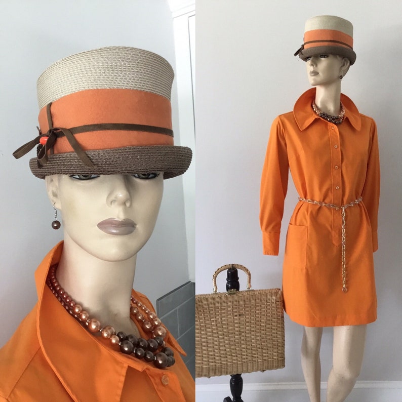Op de afbeelding: Een mannequin draagt een feloranje jurk met een bruine en beige hoed. De jurk heeft een kraag, lange mouwen en een kettingriem. De mannequin draagt ook een bruine parelketting en oorbellen.
