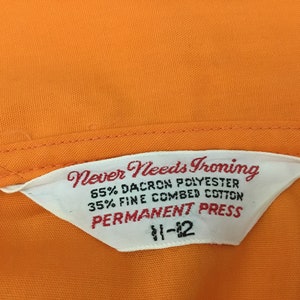 Op de afbeelding: Wit etiket met de tekst "Never Needs Ironing 65% Dacron Polyester 35% Fine Combed Cotton Permanent Press 11-12" op een oranje stoffen achtergrond.