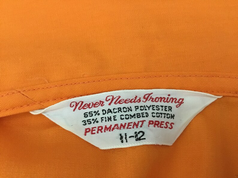 Op de afbeelding: Wit etiket met de tekst "Never Needs Ironing 65% Dacron Polyester 35% Fine Combed Cotton Permanent Press 11-12" op een oranje stoffen achtergrond.