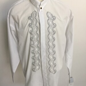 K&ouml;nnte beinhalten: Wei&szlig;es, lang&auml;rmliges Hemd mit Kn&ouml;pfen und einem Paisley-Muster auf der Vorderseite. Das Hemd hat einen Stehkragen und einen einzigen Knopfverschluss.