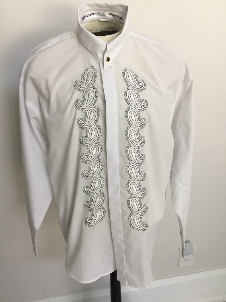 K&ouml;nnte beinhalten: Wei&szlig;es, lang&auml;rmliges Hemd mit Kn&ouml;pfen und einem Paisley-Muster auf der Vorderseite. Das Hemd hat einen Stehkragen und einen einzigen Knopfverschluss.