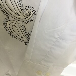 K&ouml;nnte beinhalten: Wei&szlig;es Langarmhemd mit einem schwarzen Paisley-Stickmuster auf der linken Seite des Hemdes. Das Hemd hat einen Knopfverschluss am &Auml;rmel.