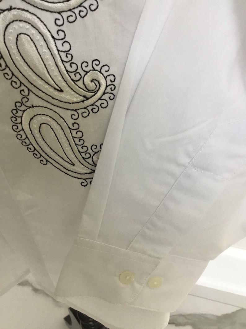 K&ouml;nnte beinhalten: Wei&szlig;es Langarmhemd mit einem schwarzen Paisley-Stickmuster auf der linken Seite des Hemdes. Das Hemd hat einen Knopfverschluss am &Auml;rmel.