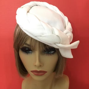 May include: A white hat with a braided design around the crown and a bow detail. The hat is made of a soft fabric and is set against a red background. This hat is a stylish accessory.