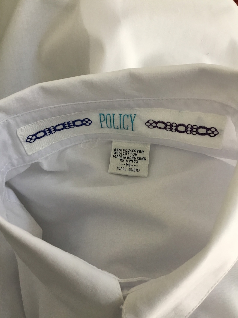 K&ouml;nnte beinhalten: Wei&szlig;es Hemd mit Kragen und einem Etikett mit der Aufschrift "POLICY" und dem Text "65% POLYESTER 35% COTTON MADE IN HONG KONG RM 57773 ---M--- (CARE OVER)"