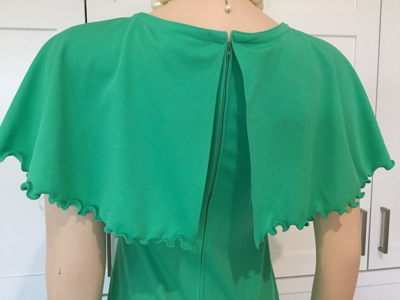 May include: A green dress with a cape-like top and a ruffled trim. The dress has a back zipper closure.