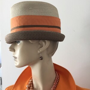 Op de afbeelding: Een strohoed met een bruine band en een oranje lint. De hoed wordt gedragen door een mannequin met een bruine parelketting en oorbellen.