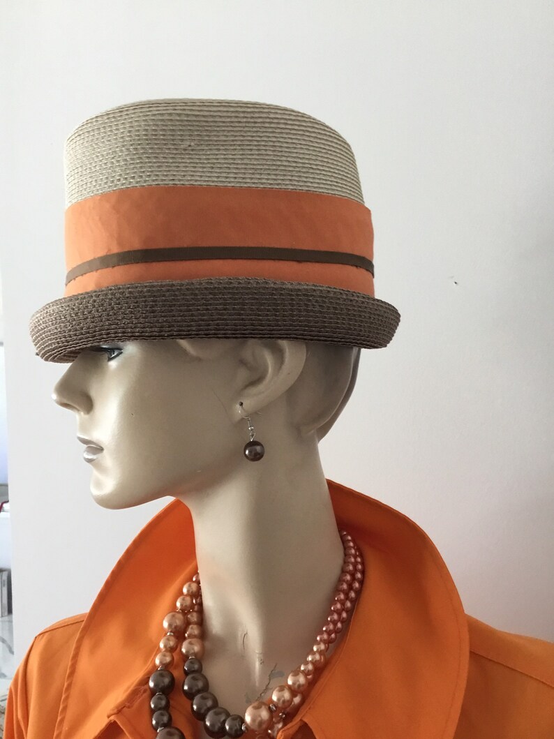 Op de afbeelding: Een strohoed met een bruine band en een oranje lint. De hoed wordt gedragen door een mannequin met een bruine parelketting en oorbellen.