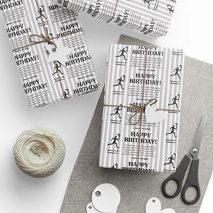 May include: Wrapped birthday gifts with gray and white striped paper, tied with twine and star-shaped tags. The paper is printed with "Happy Birthday!" and a running figure. A ball of twine, scissors, and blank tags are also visible.