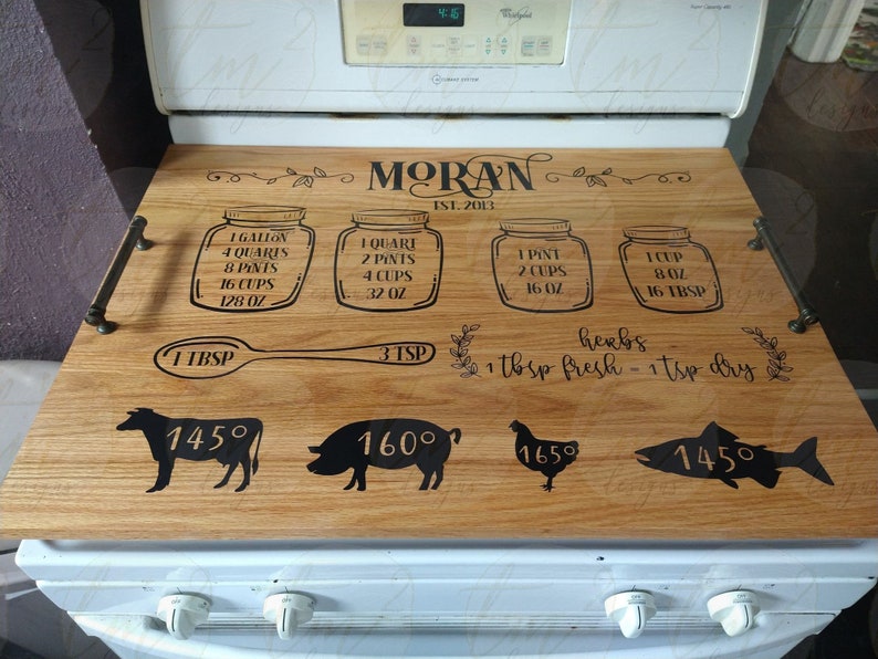 May include: A wooden stovetop tray with a personalized inscription "MORAN EST. 2013" and measurements for liquids in ounces, cups, pints, and quarts. The tray also includes a spoon, a depiction of herbs, and images of a cow, pig, chicken, and fish with corresponding temperatures.