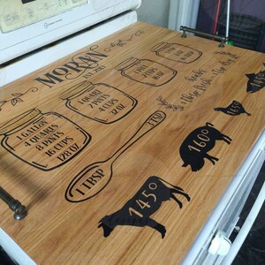 May include: A wooden stovetop cover with a black design featuring a spoon, jars with measurements, and animals. The text "MORAN EST. 2013" is printed on the cover.