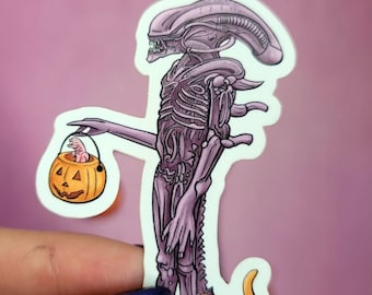 May include: A purple alien holding a jack-o-lantern with a small alien inside. A small orange cat is looking up at the alien. The sticker says "The Dawn Shine Art".